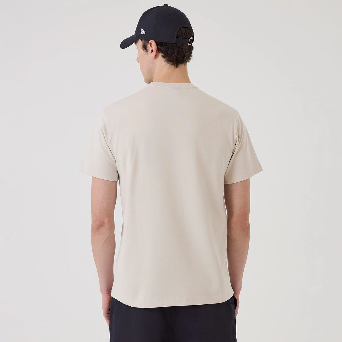 New Era SK8 Graphic Cream T-Shirt, rear view showing the plain cream back with short sleeves, worn by a model also featuring a black cap with a small embossed New Era logo on the side.