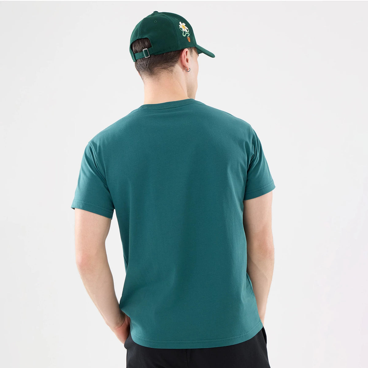 New Era Earth Day Dark Green T-Shirt, on-model view showing the back of the dark green t-shirt, paired with a matching cap that features an embroidered daisy on the side, worn by a person with their hands in pockets.