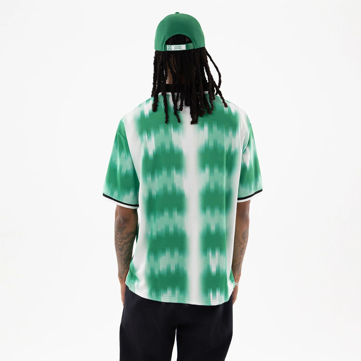 Boston Celtics NBA Green Soccer Jersey, back view on a model wearing a green cap, featuring a green and white pattern with black trim on the sleeves, displaying a relaxed fit and straight hem above dark trousers.