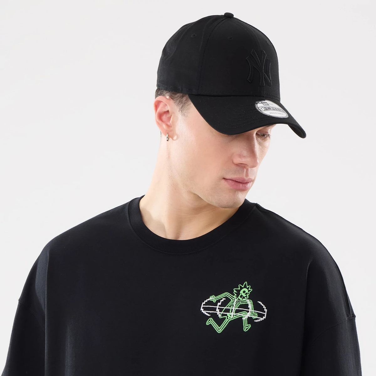 New Era Rick And Morty X-Ray Black T-Shirt, on-model view showing a black cap with a raised logo on the front and a black t-shirt with a green cartoon graphic on the left chest, worn by a person looking down.