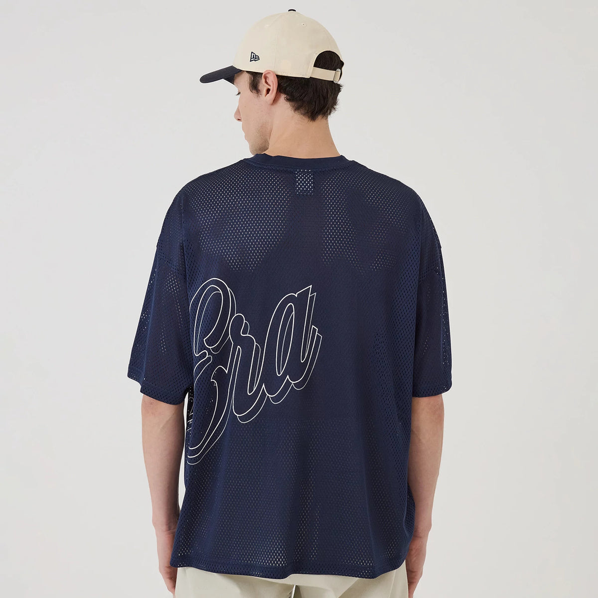 New Era Mix League Blue Oversized Mesh T-Shirt, rear view showing a large outlined "Era" text across the navy blue mesh fabric, worn by a model in a beige cap with a raised New Era logo on the side.