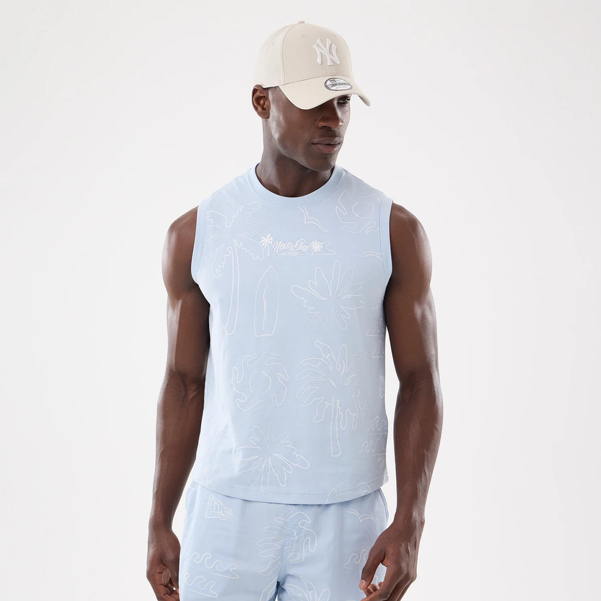 New Era Resort Blue Tank Top, front view on a model with matching shorts and a beige New York Yankees cap, featuring white palm tree embroidery on the chest and an overall tropical pattern.