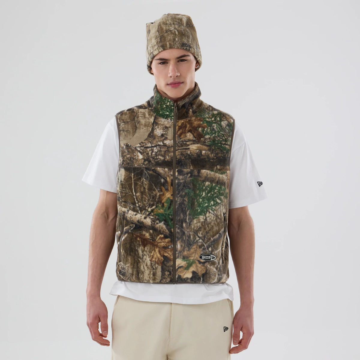 Thisisneverthat X New Era Revolution Fleece Real Tree Vest, front view on a model wearing a matching beanie and white T-shirt, showing a realistic tree camouflage pattern with an embroidered patch on the lower front.