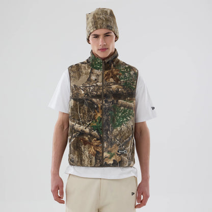 Thisisneverthat X New Era Revolution Fleece Real Tree Vest, front view on a model wearing a matching beanie and white T-shirt, showing a realistic tree camouflage pattern with an embroidered patch on the lower front.