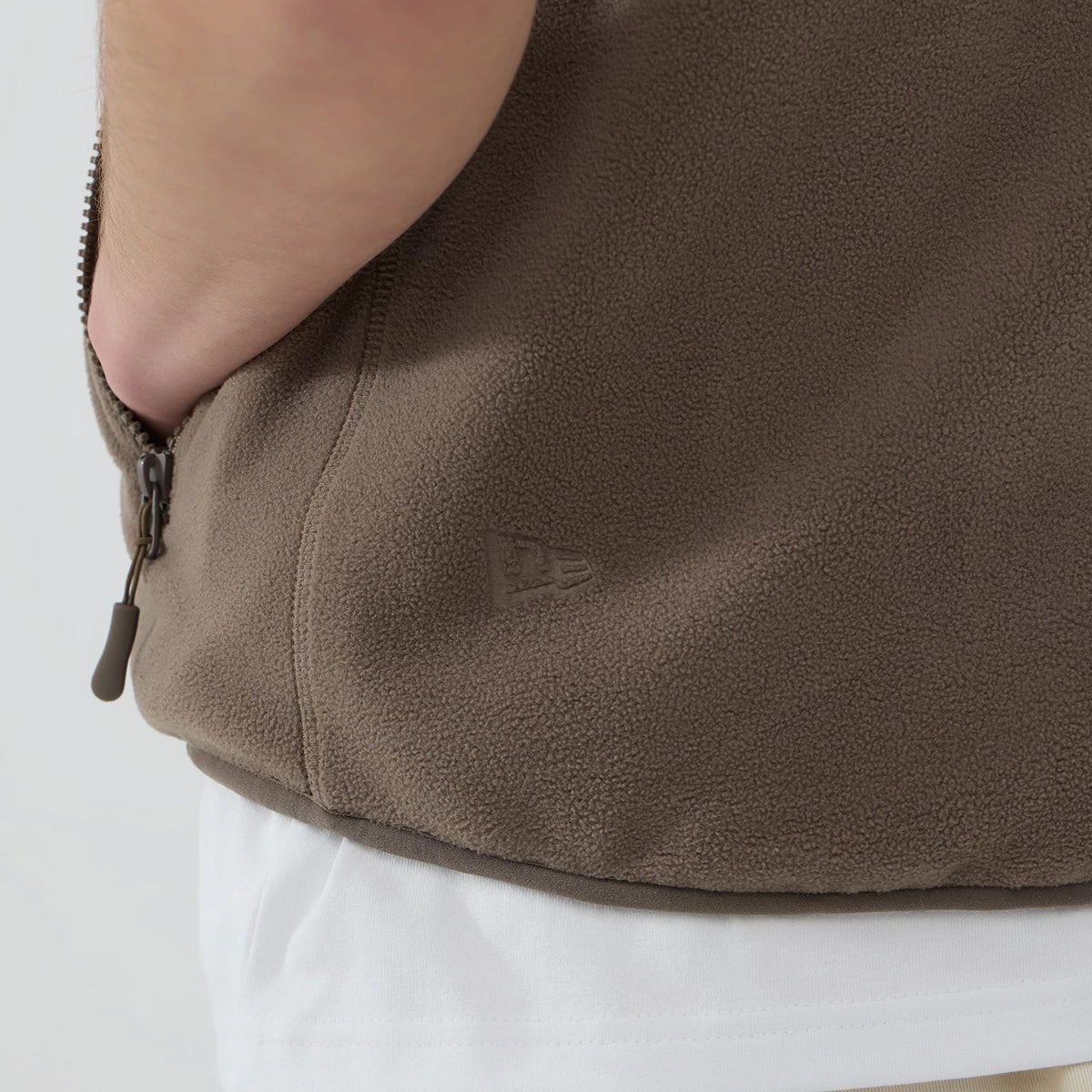 Thisisneverthat X New Era Revolution Fleece Real Tree Vest, side view on a model showing a brown fleece texture with discreet New Era flag logo stitching near the side seam above the hem of a white underlayer.