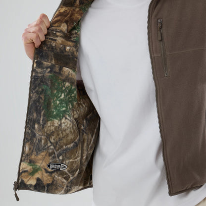 Thisisneverthat X New Era Revolution Fleece Real Tree Vest, inside view with a hand holding the open side, showing the Real Tree camo pattern interior and a New Era patch near the pocket.