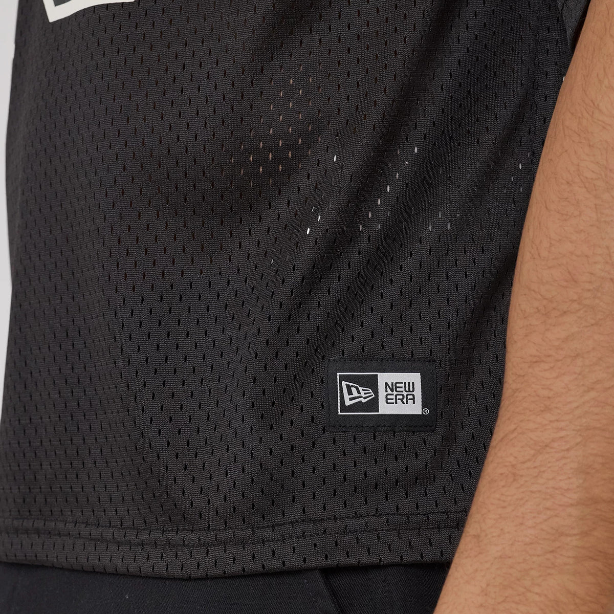 New Era Mesh Football Black Short Sleeve Shirt, side view showing the mesh texture and a New Era flag logo patch near the hem, worn by a model with part of the arm visible.