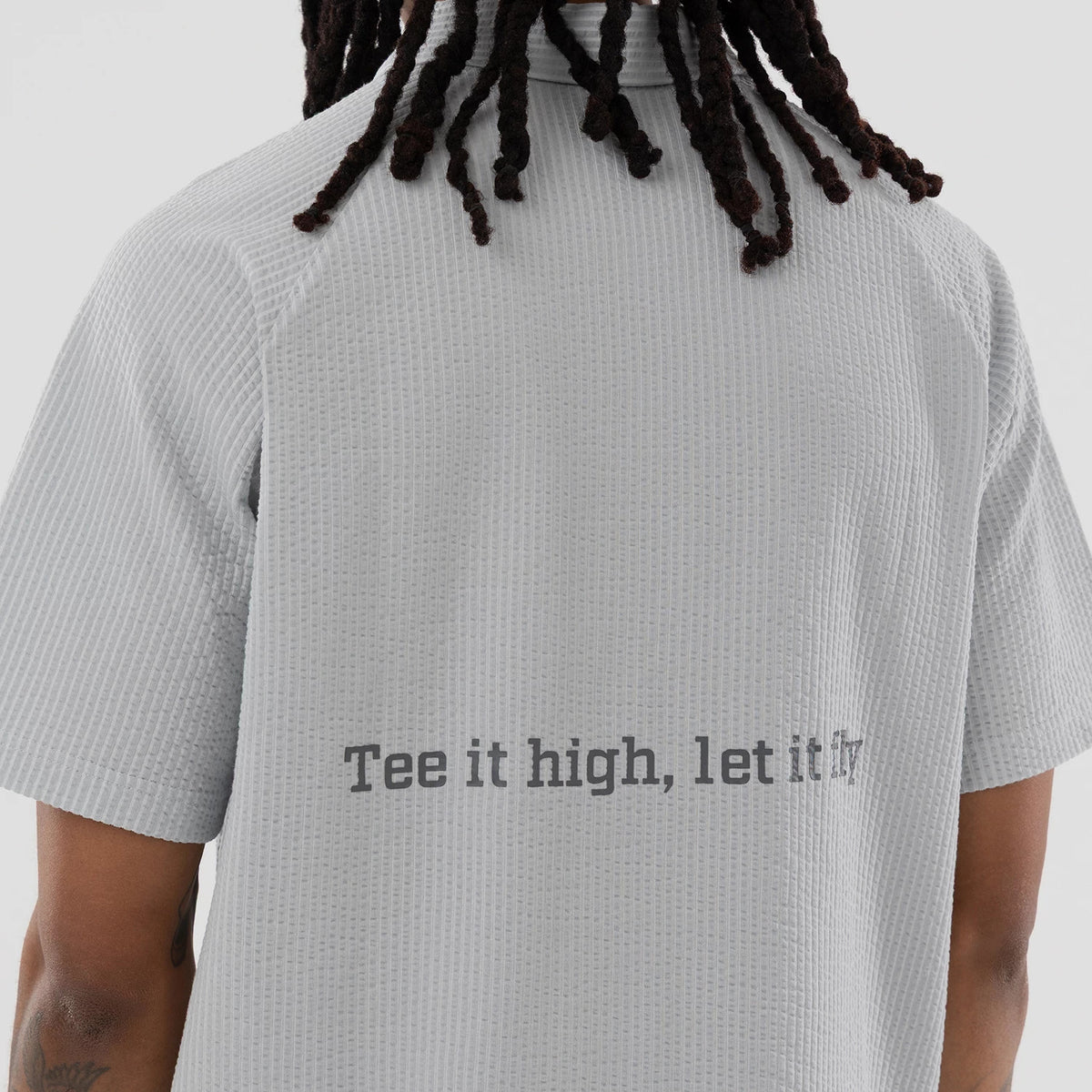 New Era Seersucker Grey Polo T-Shirt, rear view showing the textured grey fabric with short sleeves and the phrase "Tee it high, let it fly" printed in dark grey across the back above the lower hem.