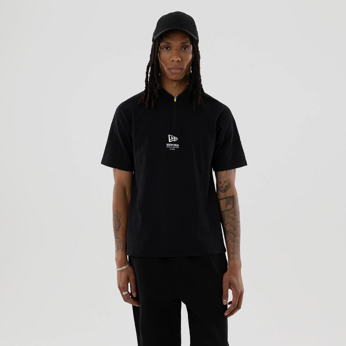 New Era Seersucker Black Polo T-Shirt, front view showing the black short-sleeve polo with a raised New Era logo on the chest, a high collar with a yellow zip pull, worn by a person also wearing a black cap.