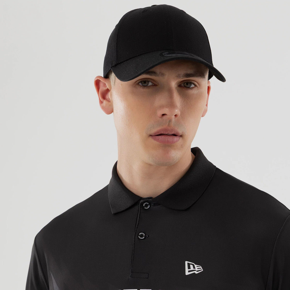 New Era Grid Black Polo T-Shirt, on-model view featuring a black short-sleeve style with a three-button placket, a light grey raised New Era logo on the left chest, and a black cap with a debossed logo on the front.