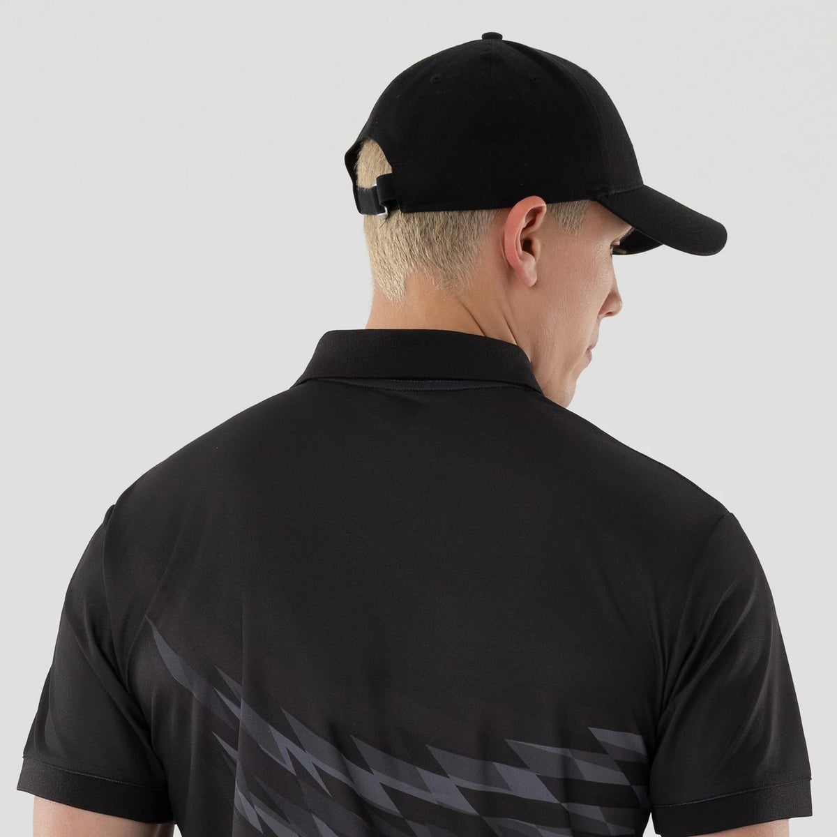 New Era Grid Black Polo T-Shirt, rear view showing a black polo shirt with a subtle grid pattern and short sleeves, worn by a person with a black cap featuring a debossed New Era logo on the back near the adjuster.