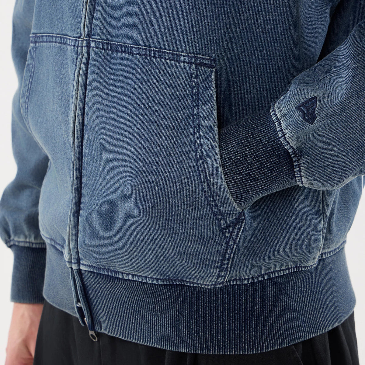 New Era Washed Blue Full Zip Hoodie, close-up showing the front pocket detail on the faded blue fabric, ribbed hem and cuffs, with a dark embroidered logo on the sleeve above the wrist.