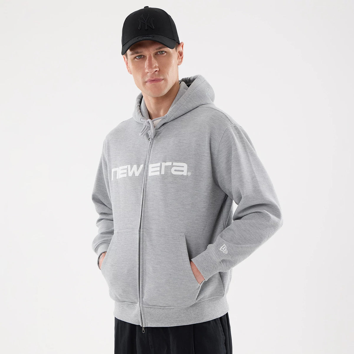 New Era Waffle Grey Full Zip Hoodie, front view showing the grey hoodie with a white printed "New Era" logo across the chest, a full-length zip, two front pockets, and an embossed black logo on a cap worn by the model.