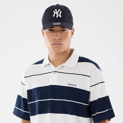 New Era Stripe White Polo T-Shirt, on-model view showing the white polo with navy stripes, a raised New Era logo on the left chest, paired with a black cap featuring an embossed white logo.