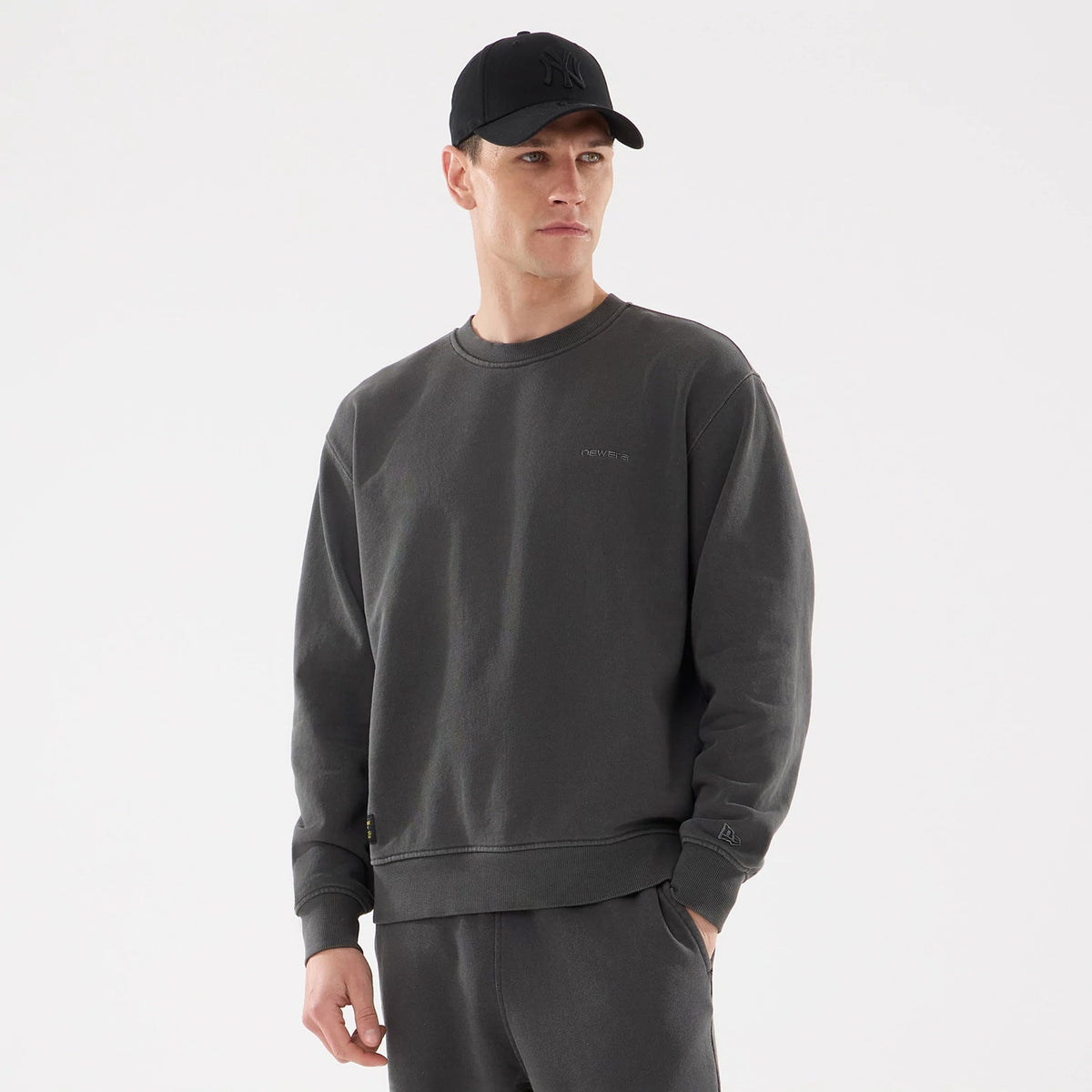 New Era Originators Black Crew Neck Sweater, on-model view with matching black cap, showing subtle front logo embroidery, ribbed cuffs and hem, and New Era flag logo on the left sleeve.