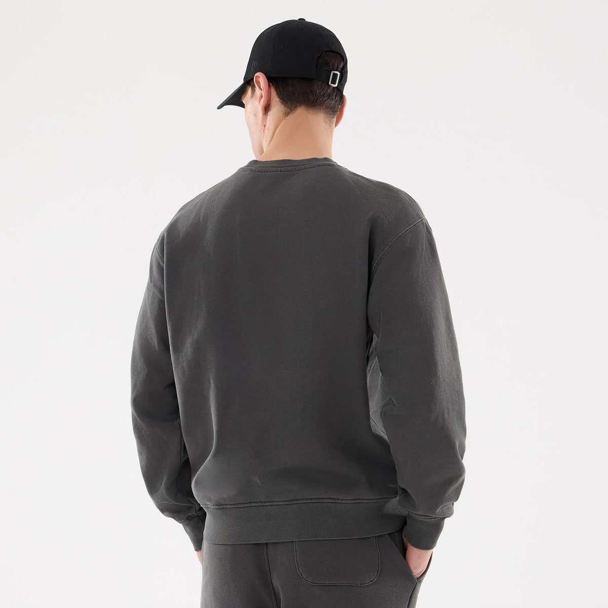 New Era Originators Black Crew Neck Sweater, rear view on a model dressed in matching black trousers and a black cap, showing the plain back design and relaxed fit with ribbed cuffs and hemline.