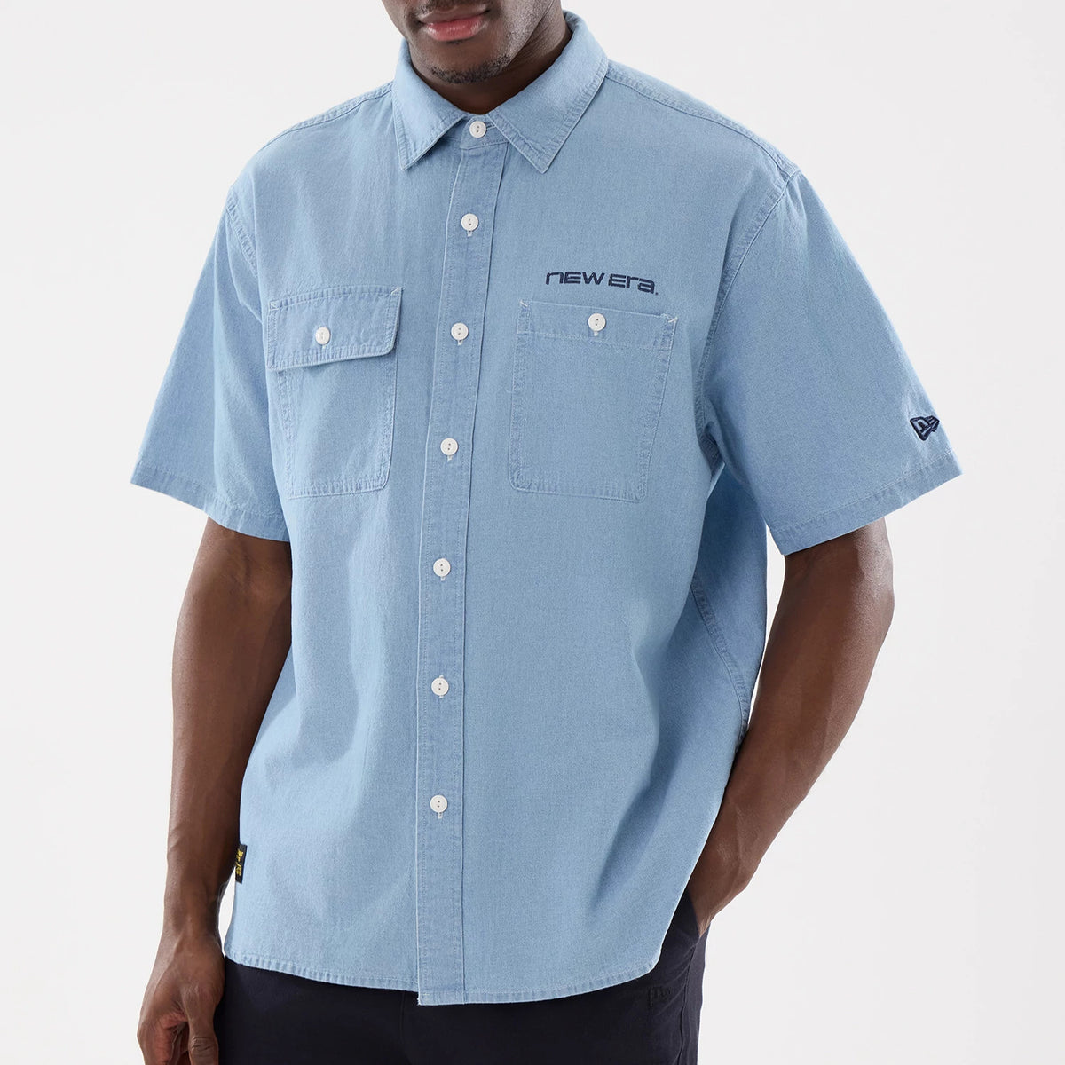 New Era Originators Chambray Bright Blue Short Sleeve Shirt, front view on a model with two buttoned chest pockets, embroidered New Era logo on the right chest, and New Era flag logo on the left sleeve.