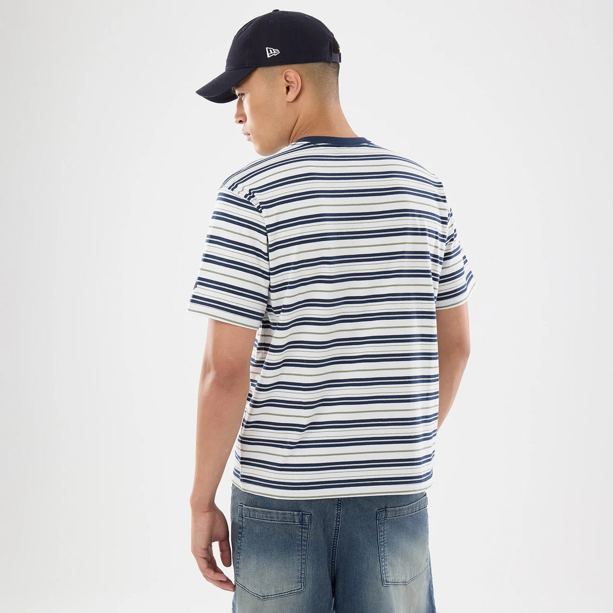 New Era Boarder Stripe White T-Shirt, on-model view showing the back with navy and white horizontal stripes and a plain round neckline, paired with denim shorts and a navy cap featuring a raised logo on the side.