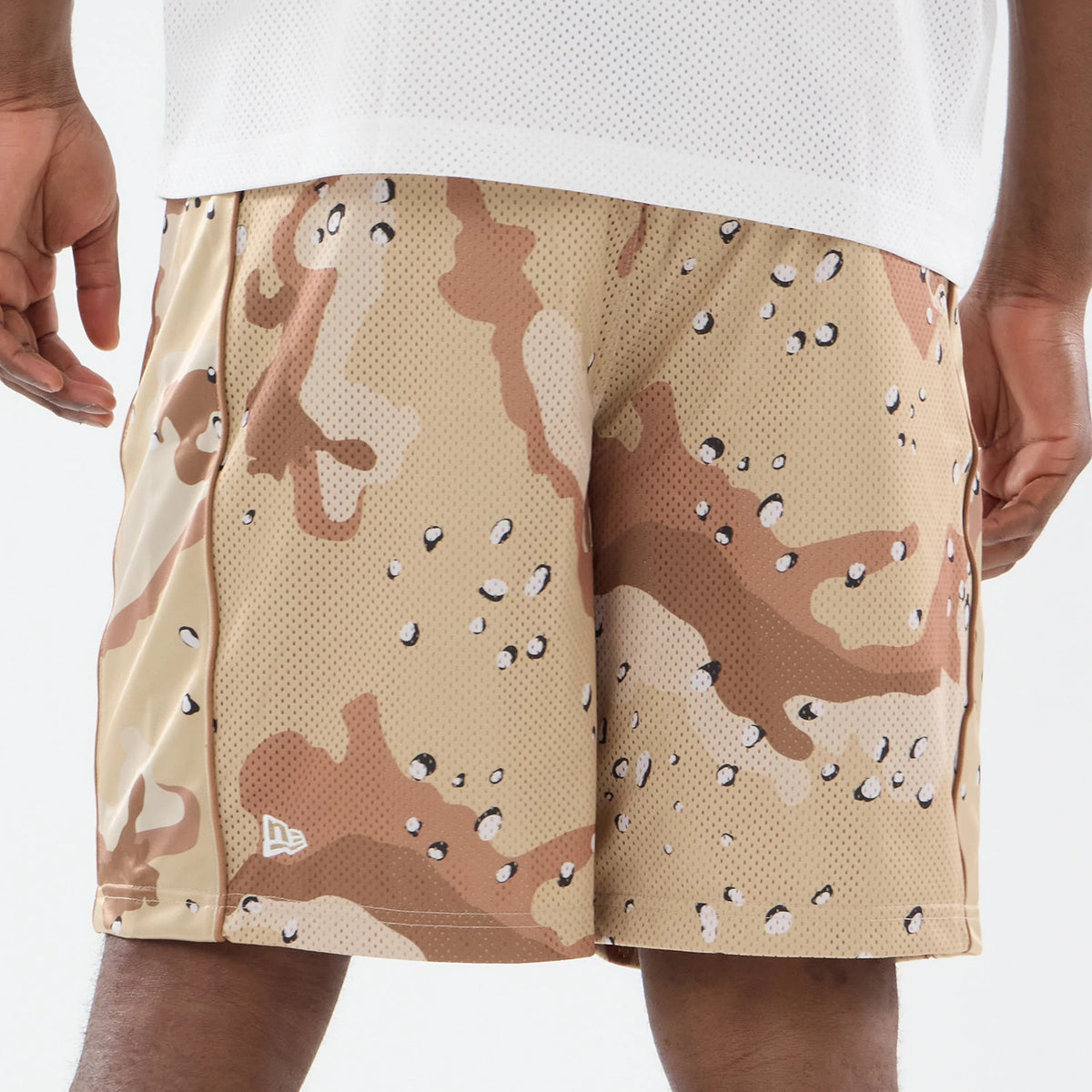 New Era Bermuda Woodland Camo Oversized Shorts, on-model view showing the beige and brown camo pattern across the shorts, with a raised New Era logo on the left leg near the hem and a relaxed fit reaching above the knees.