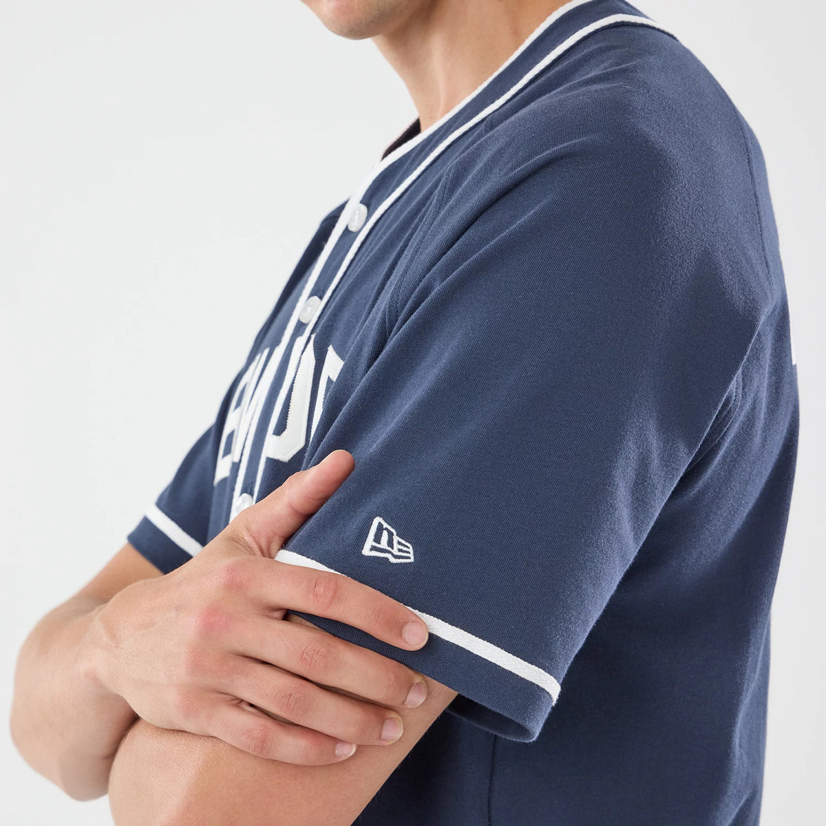 New Era New York Navy Jersey, close-up on a model showing the white New Era flag logo on the sleeve and white piping along the collar and button placket, with the model's arms crossed.