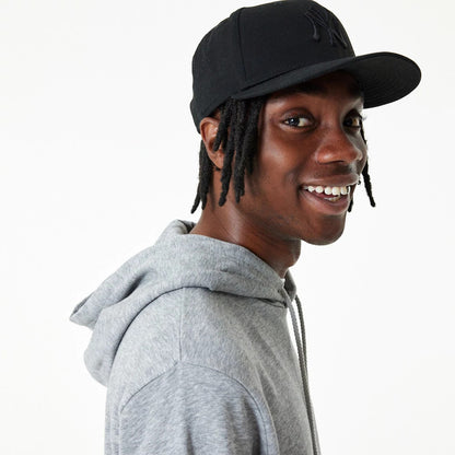New Era Essential Grey Pullover Hoodie, on-model view showing a smiling individual wearing the grey hoodie with the hood down, paired with a black cap featuring a raised logo on the front.