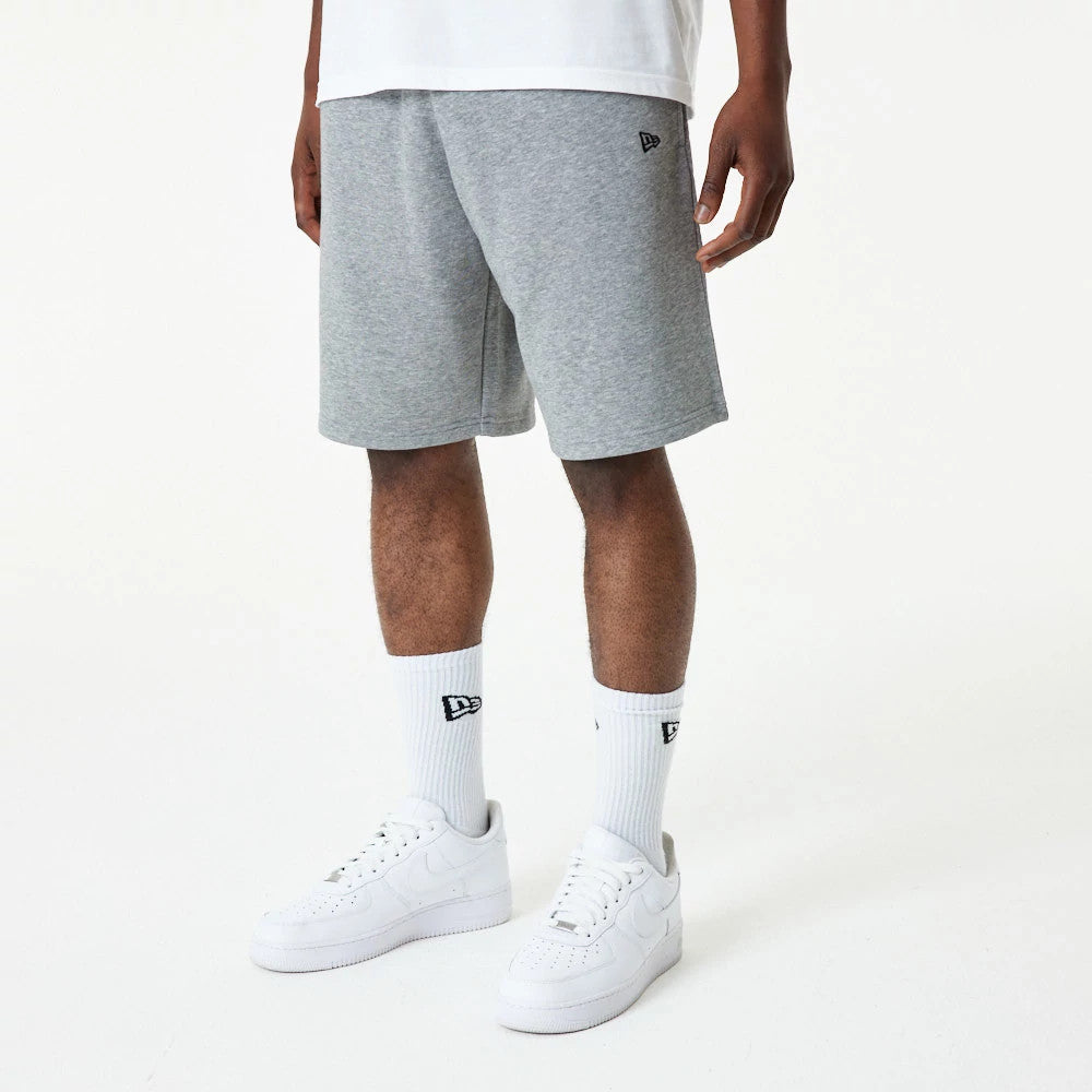 New Era Essential Grey Shorts, on-model view showing grey knee-length shorts with an embroidered New Era logo on the left leg, paired with white socks featuring a black New Era logo and white trainers.