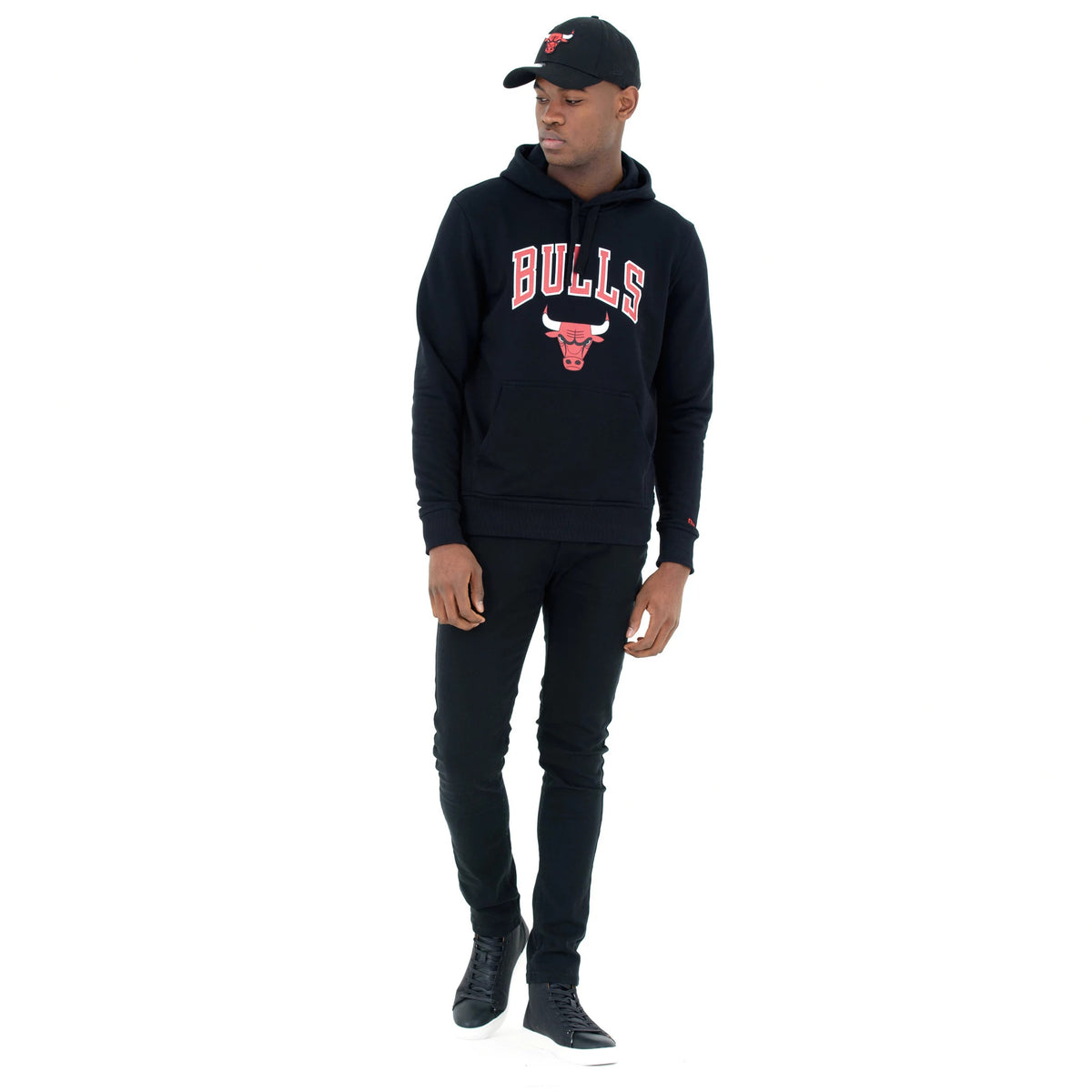 Chicago Bulls NBA Regular Black Pullover Hoodie, on-model view showing the black hoodie with bold red BULLS lettering and a bull logo on the chest, paired with black trousers and a black cap with an embroidered red bull logo.
