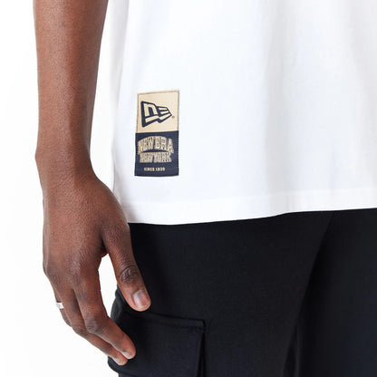 New Era Sticker White Oversized T-Shirt, side view showing a white T-shirt with a large rectangular patch near the hem featuring a debossed New Era logo and text, worn over black trousers with a hand resting at the side.