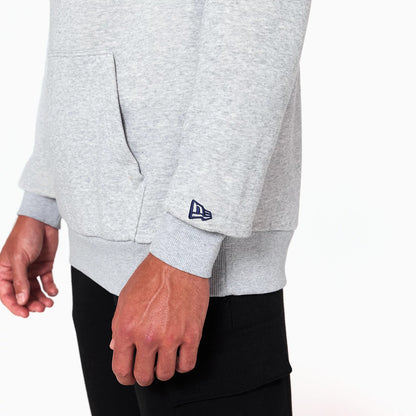 New Era Washington Wizards NBA Grey Pullover Hoodie, side view showing the light grey fabric with a front pocket and a raised dark blue New Era logo on the left sleeve cuff, part of a casual outfit with black trousers.