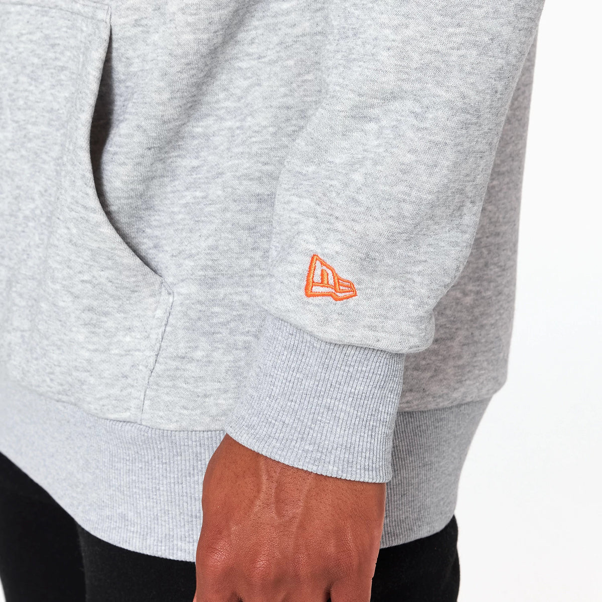 New Era New York Knicks NBA Grey Pullover Hoodie, side view showing the light grey fabric with a raised orange New Era logo on the left cuff above the ribbed hem and a large front pocket beside it.