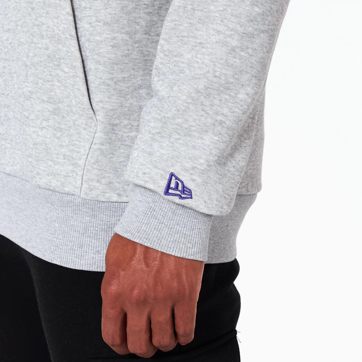 New Era Charlotte Hornets NBA Grey Pullover Hoodie, side view showing the light grey fabric with a ribbed cuff on the sleeve, a raised purple New Era logo near the wrist above the casual black trousers worn by the model.