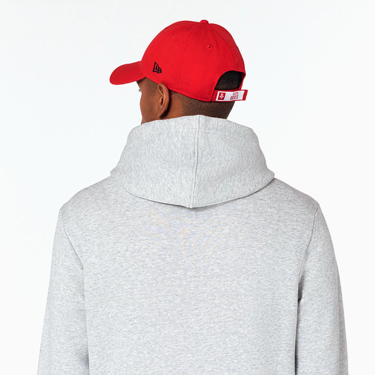 New Era Houston Rockets NBA Grey Pullover Hoodie, rear view showing the light grey fabric and large hood, worn by a model also wearing a red cap with a raised New Era logo on the side and adjustable strap at the back.