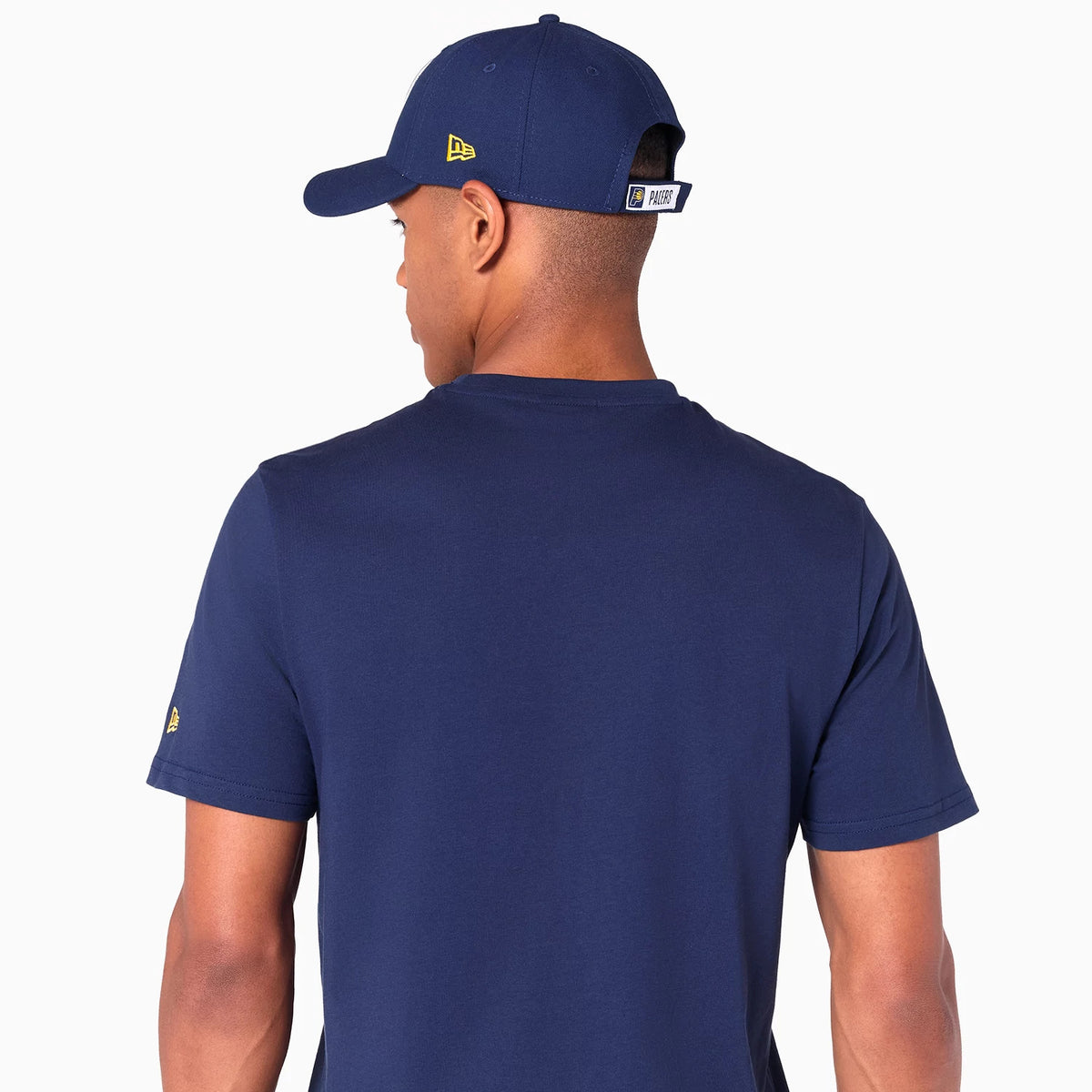 New Era Indiana Pacers NBA Graphic Navy T-Shirt, on-model view showing the back of the navy T-shirt with the sleeves reaching mid-arm, a yellow embroidered New Era logo on the left sleeve, paired with a navy cap with a yellow New Era logo.
