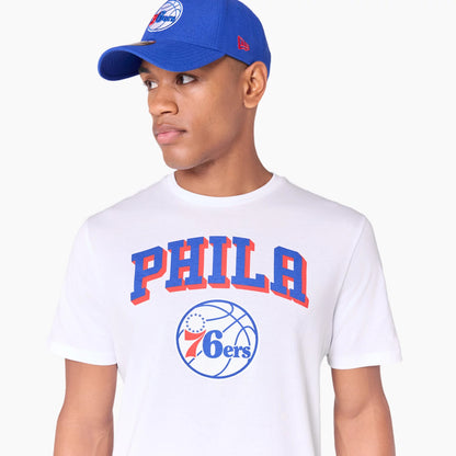 New Era Philadelphia 76ers NBA White T-Shirt, on-model view showing the blue and red PHILA text across the chest above a detailed basketball graphic with a 76ers logo, paired with a blue cap featuring a raised New Era logo on the side.