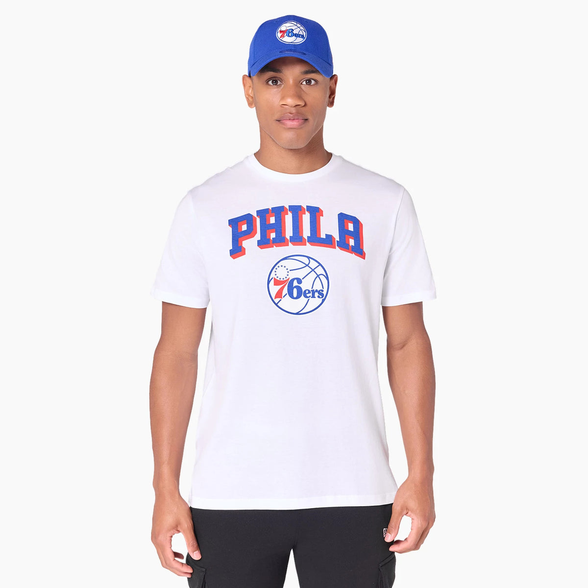 New Era Philadelphia 76ers NBA White T-Shirt, on-model view showing the white shirt with large red and blue PHILA lettering across the chest above a debossed 76ers logo in blue and red, paired with a blue cap featuring the same logo.