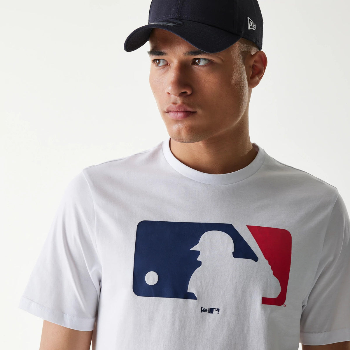 New Era MLB Generic Logo MLB Regular White T-Shirt, on-model view showing a large blue and red MLB logo printed on the chest, with a black cap featuring a raised New Era logo on the side worn by the model.