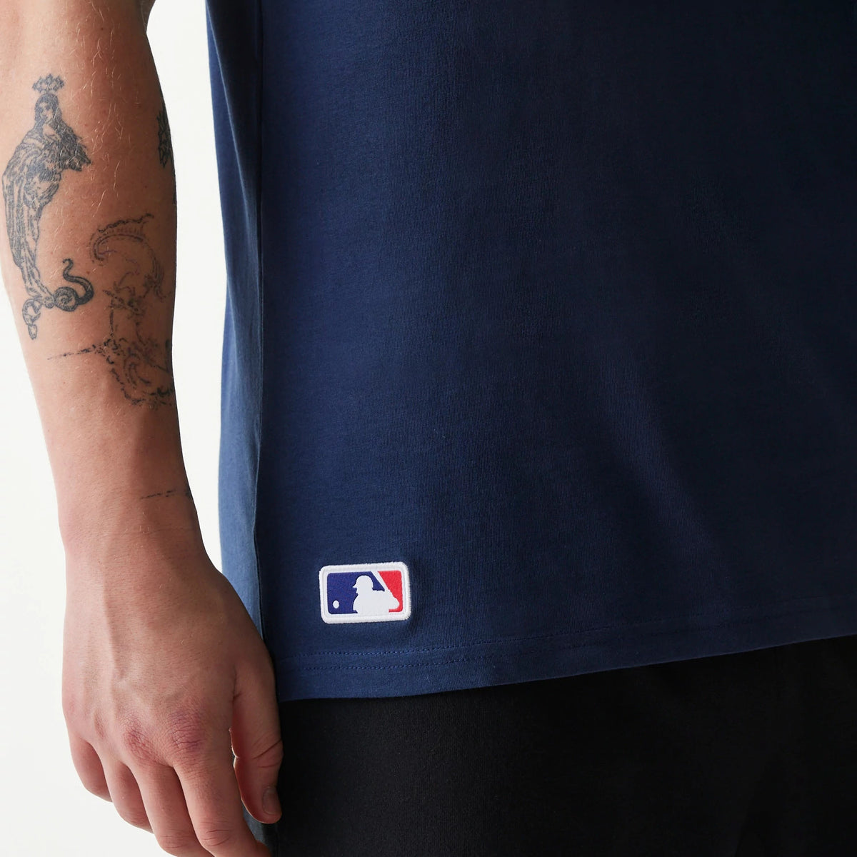 New Era Texas Rangers MLB Regular Dark Blue T-Shirt, side view showing the bottom half with the white embroidered MLB logo on the lower left hem and the dark blue fabric, partially covering the wearer’s arm with tattoos.