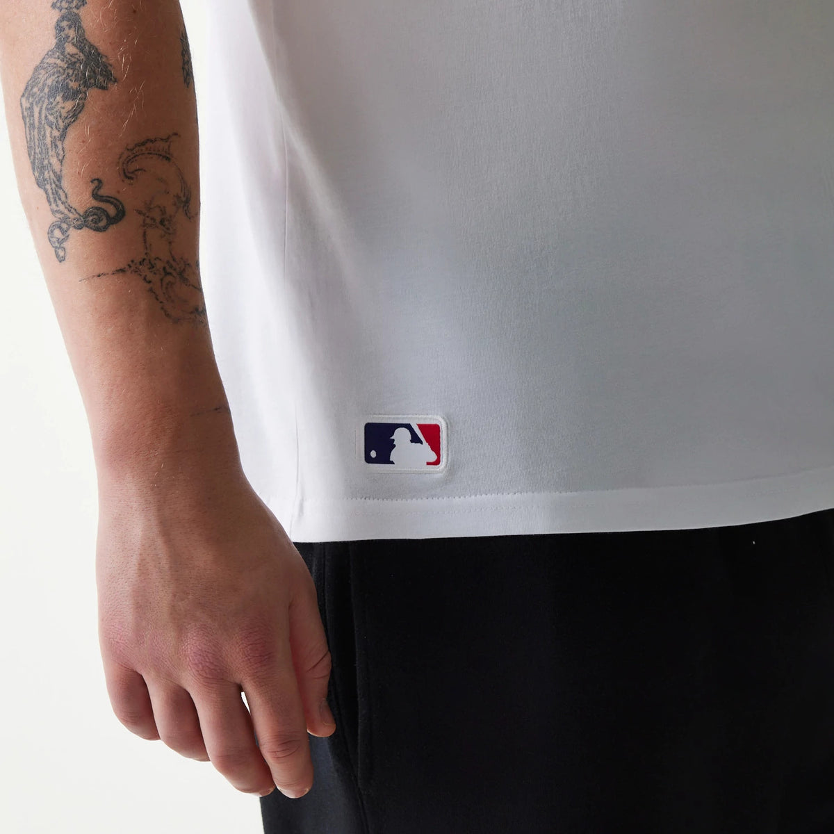 New Era Tampa Bay Rays MLB Regular White T-Shirt, side view showing the lower edge of the white fabric with an embroidered MLB logo patch near the hem on the left side, next to a tattooed arm.