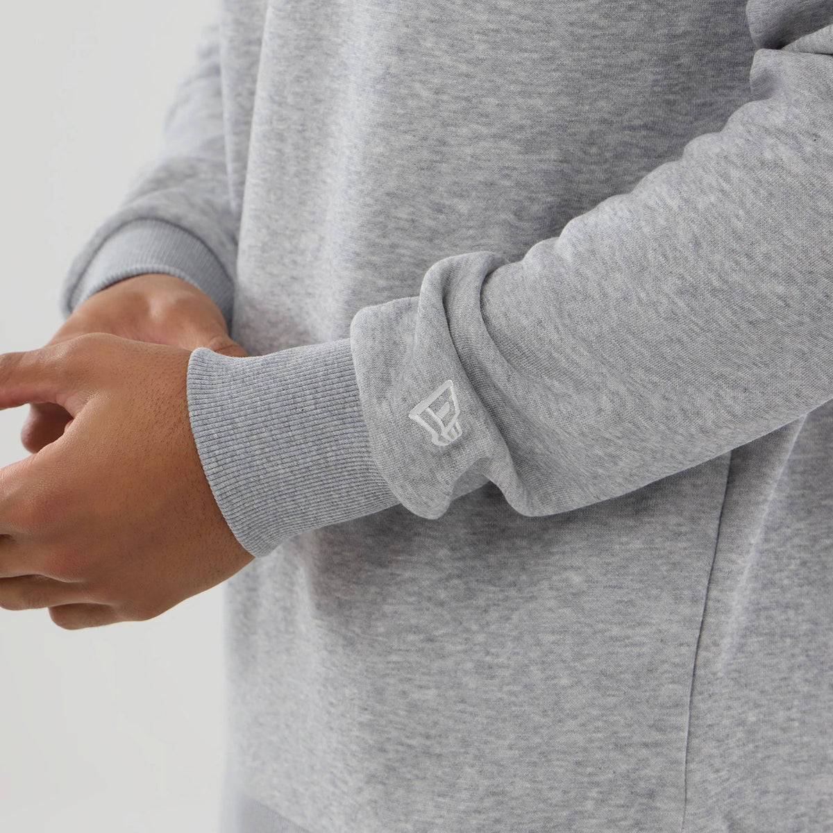New Era MLB Generic Logo Grey Crew Neck Sweater, close-up showing the ribbed cuff with a white New Era flag logo on the left sleeve, highlighting the soft grey fabric texture and relaxed fit on a model's arm.
