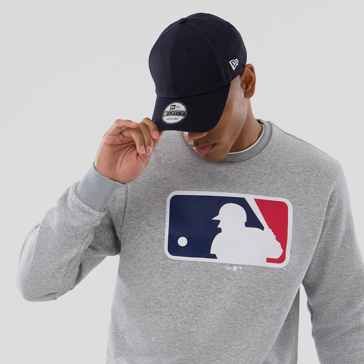 New Era MLB Generic Logo Grey Crew Neck Sweater, on-model view wearing a navy New Era 9FORTY cap, showcasing the large colourful MLB logo on the chest and the New Era flag logo on the left side of the cap.