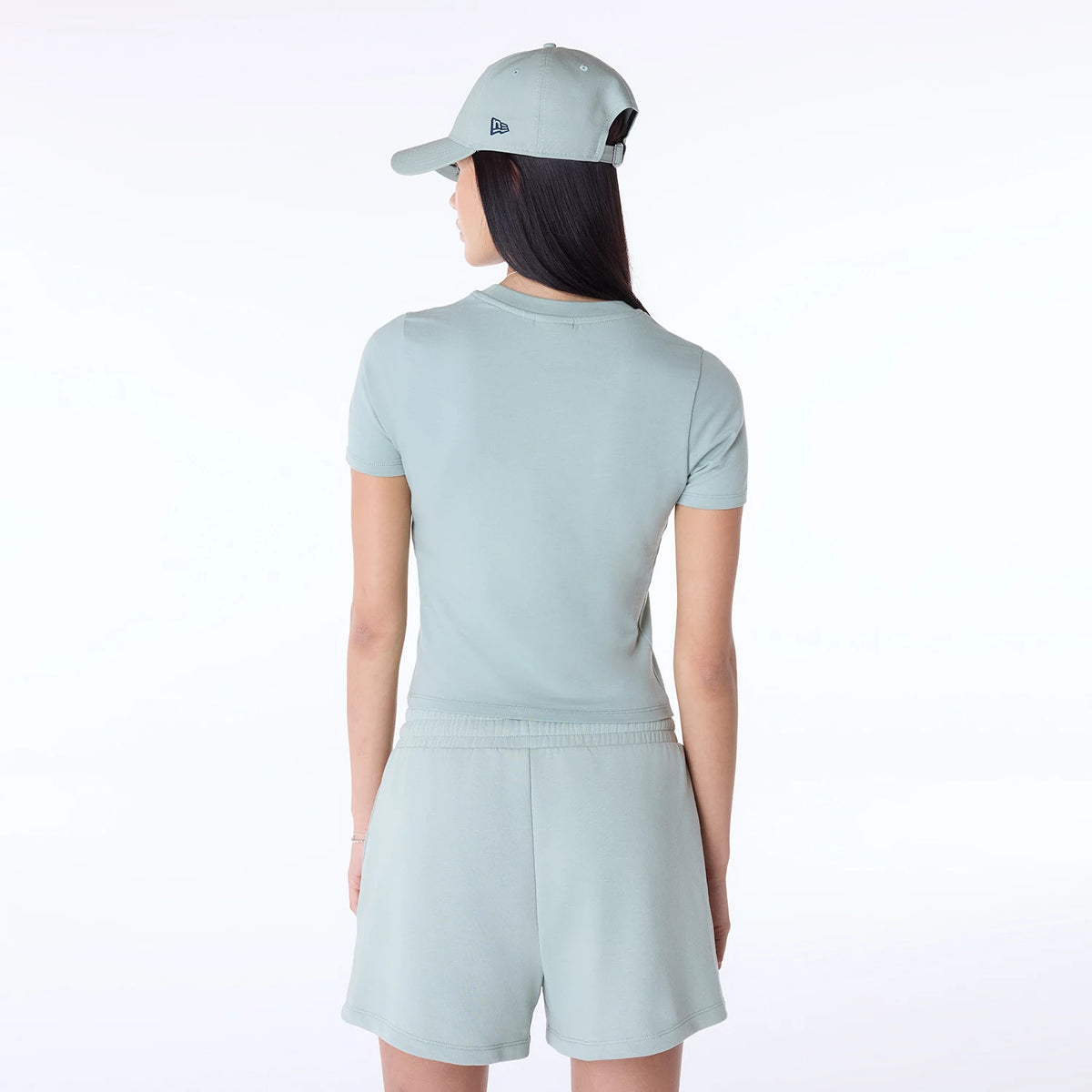 Womens New Era Slim Pastel Blue T-Shirt, on-model rear view showcasing the fitted pastel blue shirt paired with matching shorts, a blue debossed New Era logo on a pastel blue cap, and the model's long dark hair cascading down.