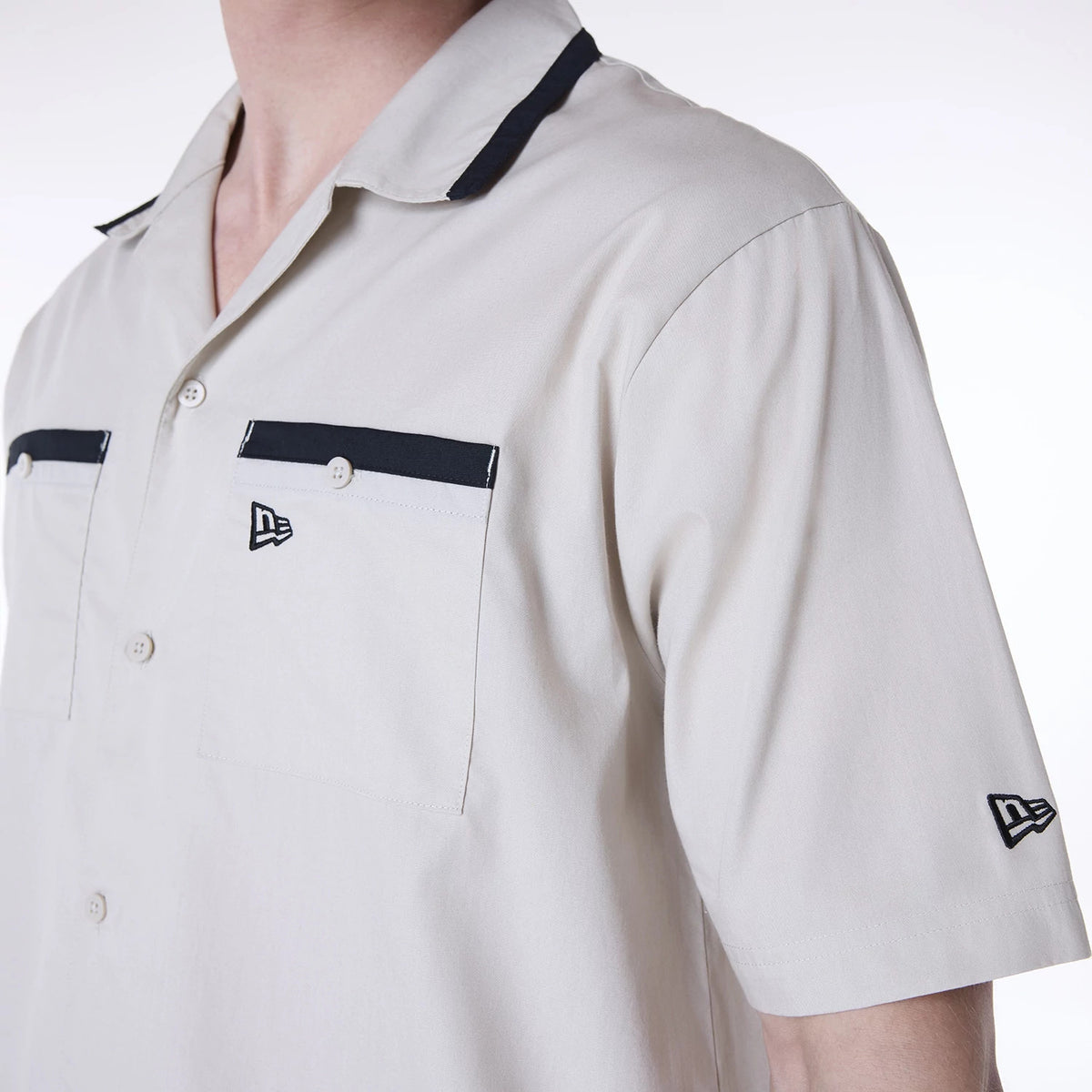 New Era Revere Cream Short Sleeve Shirt, three-quarter view on a model showing contrasting black trim on the collar and pocket edges, white buttons, and New Era flag logos on the chest pocket and left sleeve.
