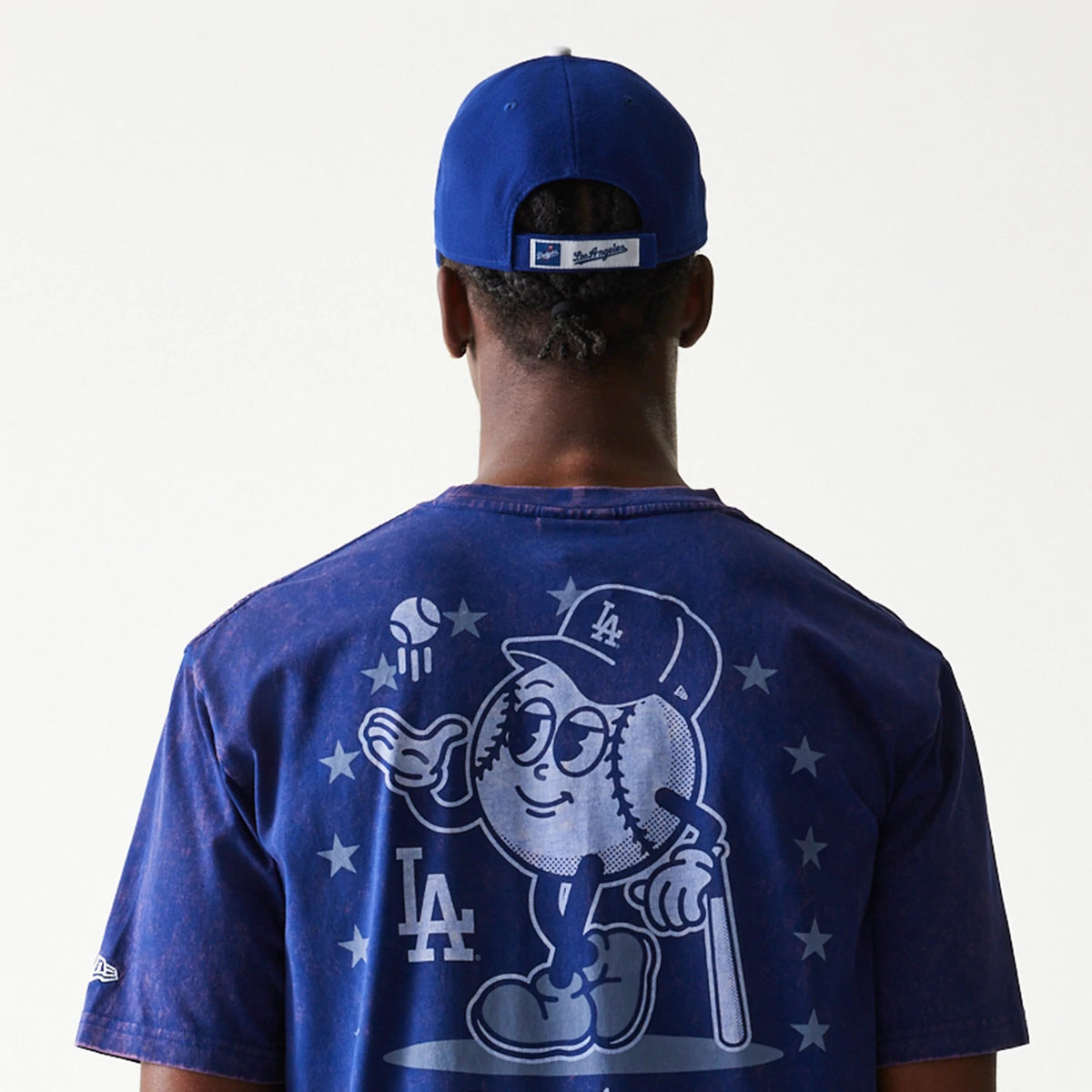 New Era LA Dodgers MLB Character Dark Blue T-Shirt, rear view showing a graphic of a baseball character wearing an LA cap with stars around, the LA logo below, and a blue cap with a debossed New Era logo worn by the model.