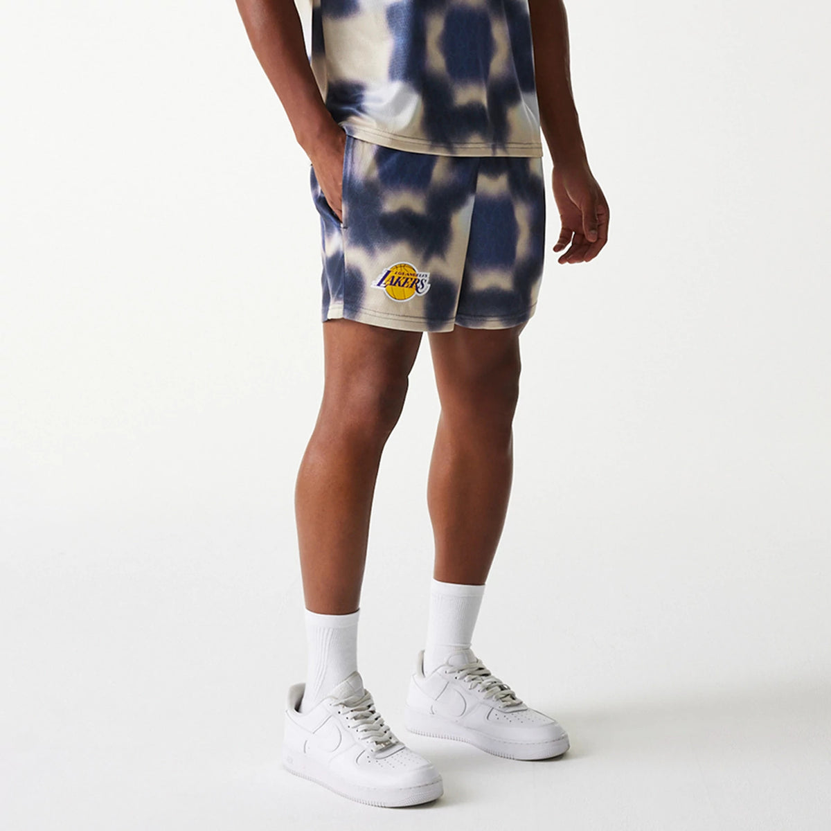 New Era LA Lakers NBA All Over Print Cream Shorts, on-model view showing cream shorts with a blue tie-dye pattern, the yellow and purple Lakers logo printed on the left leg near the hem, paired with white trainers and socks.