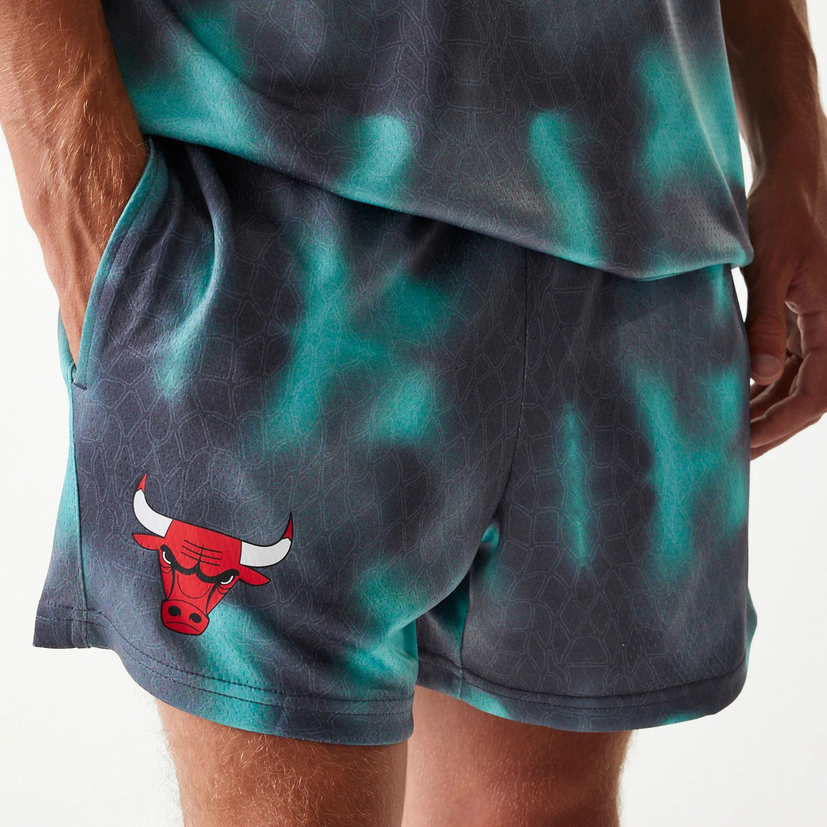 New Era Chicago Bulls NBA All Over Print Green Shorts, front view showing a vivid green pattern on dark fabric with the red embossed Chicago Bulls logo on the left leg, a hand partially in the side pocket, and the hem resting above the knee.