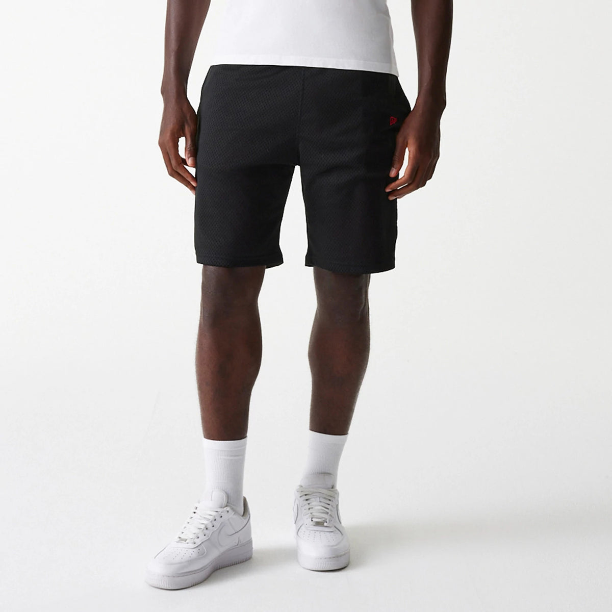 New Era Black Mesh Shorts, front view showing the knee-length black shorts with a raised red New Era logo on the left leg, hands relaxed by the sides, paired with white socks and trainers.