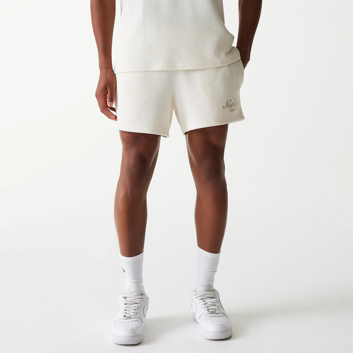 New Era Waffle Open White Shorts, front view showing white shorts with a debossed New Era logo on the left thigh, worn with white socks and trainers, part of a matching waffle-textured set.