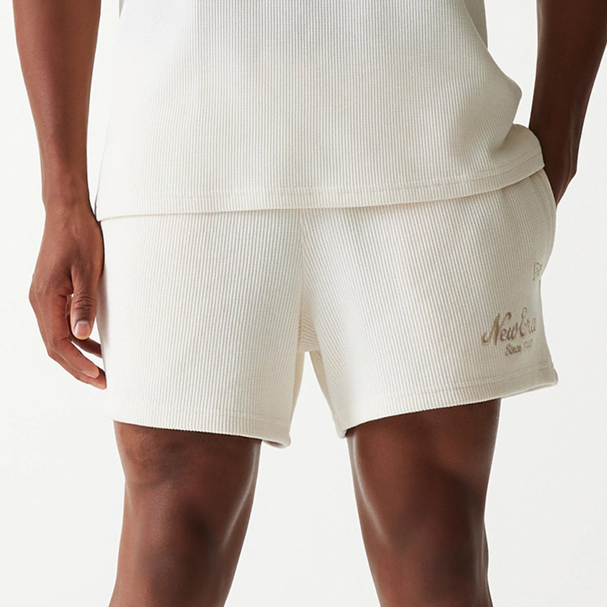 New Era Waffle Open White Shorts, front view showing the ribbed white fabric with a raised New Era logo in beige above the left hem, paired with a matching top, highlighting the casual and relaxed fit.