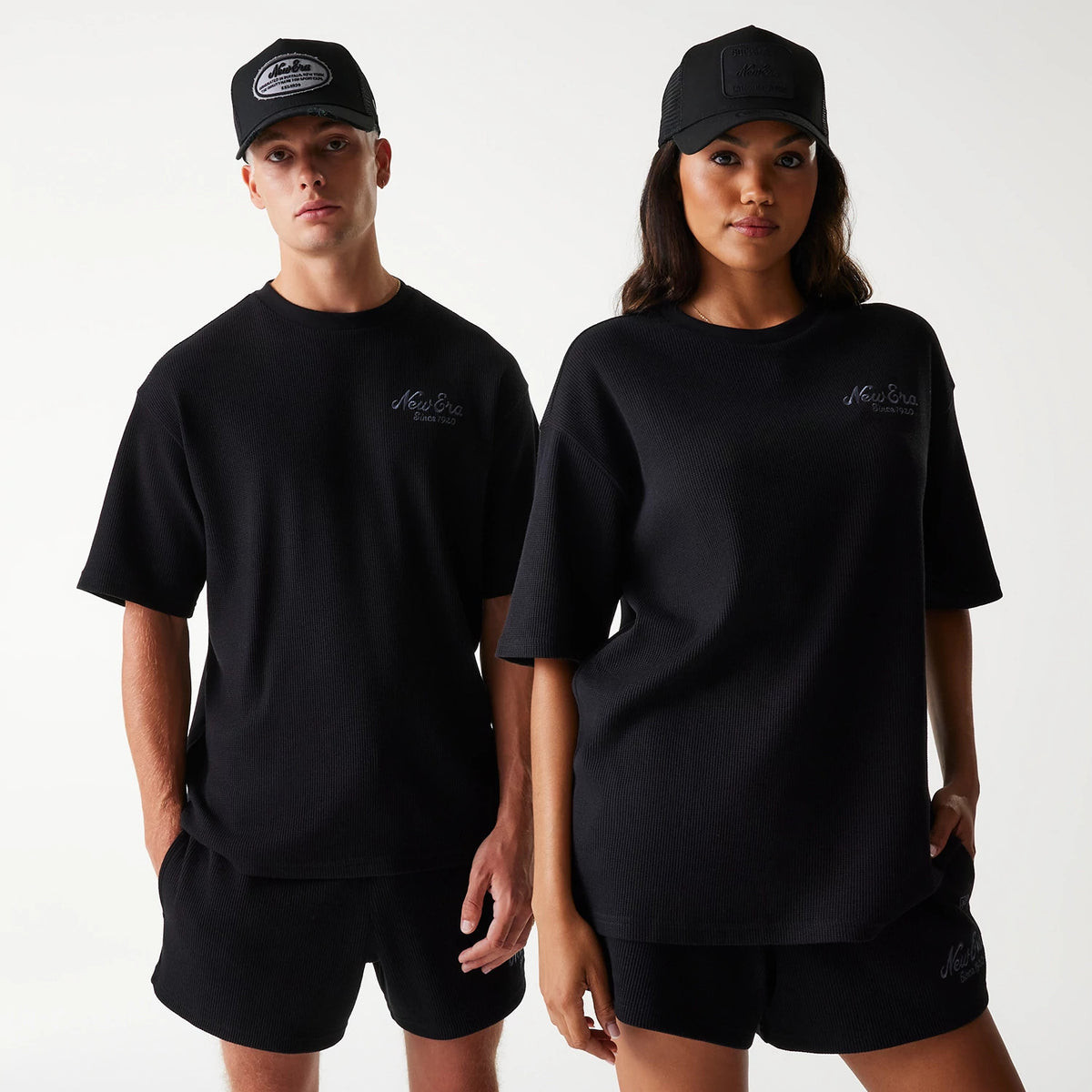 New Era Waffle Black Oversized T-Shirt, on-model view showing two models wearing black oversized T-shirts with short sleeves, accompanied by black caps, each T-shirt with a debossed New Era logo on the left chest.