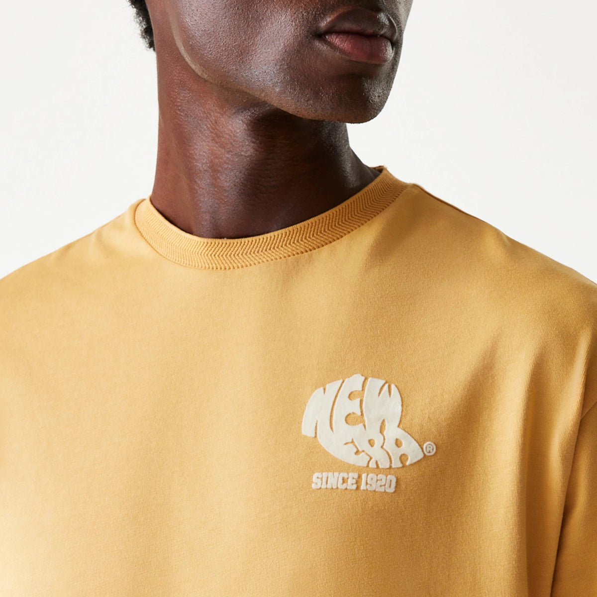 New Era Graphic Beige Oversized T-Shirt, front view showing a beige t-shirt with a ribbed round neckline and an embossed white New Era logo with 'Since 1920' text printed on the left chest.